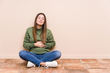 Young caucasian woman sitting on the floor isolated touches tummy, smiles gently, eating and satisfaction concept.