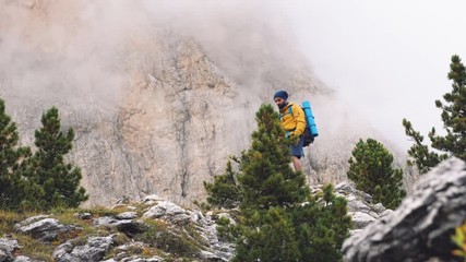 Wall Mural - young man hiker hiking on stone mountain with cloudy sky and fog. yellow jacket, backpack, black bea