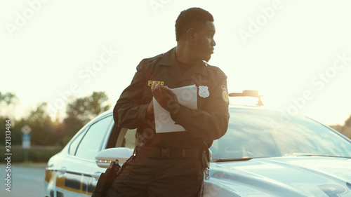 Portrait of young black police officer writing notes at accident investigation. Close-up afro cop doing paperwork turning on the car crash scene with police on background.