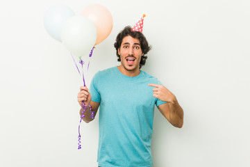 Wall Mural - Young man celebrating a party holding balloons surprised pointing at himself, smiling broadly.