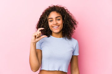 Wall Mural - Young african american woman against a pink background holding something little with forefingers, smiling and confident.