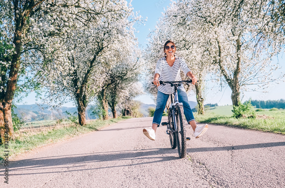 Happy smiling woman rides a bicycle on the country road under the apple blossom trees - obrazy, fototapety, plakaty 
