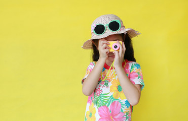 Asian little girl taking photo by toy camera. Child tourists in floral pattern summer dress and hat with sunglasses isolated on yellow background.