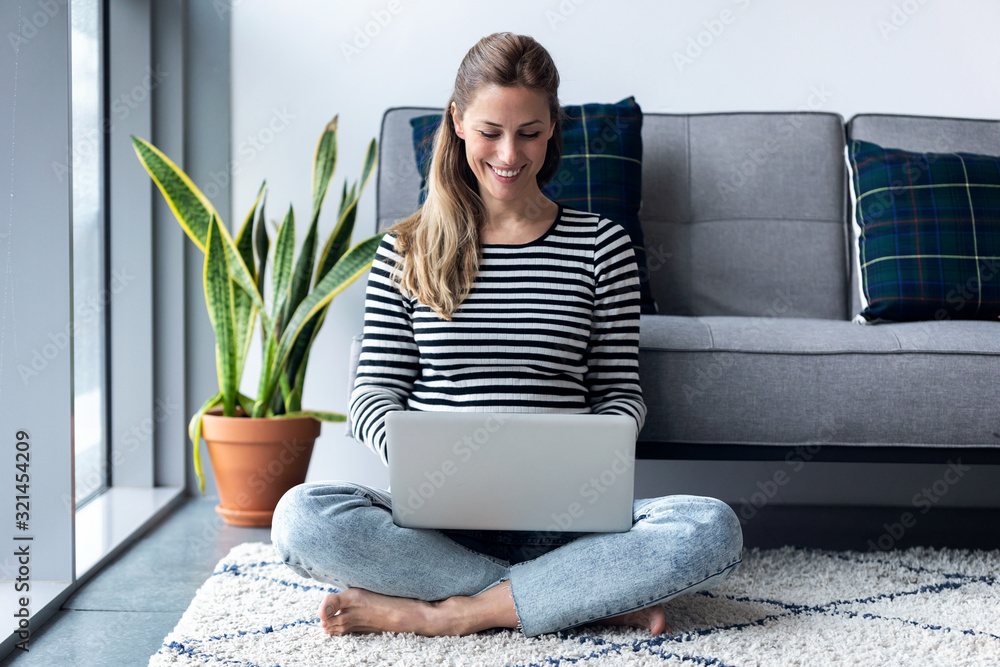 Pretty young woman using her laptop while sitting on the floor at home ...