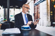 © BullRun - Aged businessman in glasses sitting and texting on tablet