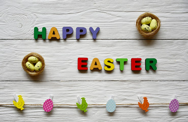 Multi-colored wooden letters making up the words Happy Easter, two nests with decorative eggs and multi-colored Easter clothespins on a white wooden background. Top view. Bright spring background. 