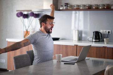 young adult man with beard and tattoo yawns, stretching in front of laptop on table with cup of coffee  and cell phone. lifestyle, freelance, social media concept