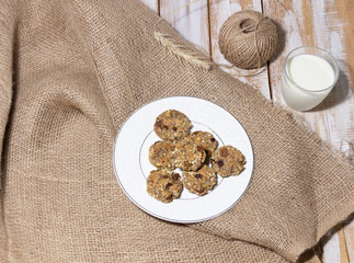 Wall Mural - Oat vegan healthy cookies baked with banana, cereals and raisin in white plate  on sackcloth on wooden background, rope, glass of milk