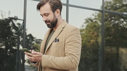 Wall Mural - Side view of Smiling pensive elegant bearded man in coat using smartphone and looking away while walking outdoors