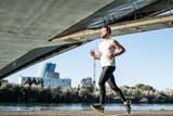 Fototapeta Przeznaczenie - young man running in a white shirt across a bridge. He's listening to music and he's got some helmets on.
