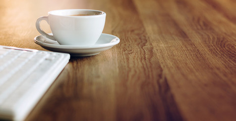 Close up of white coffee cup with out of focus computer keyboard on wooden desk