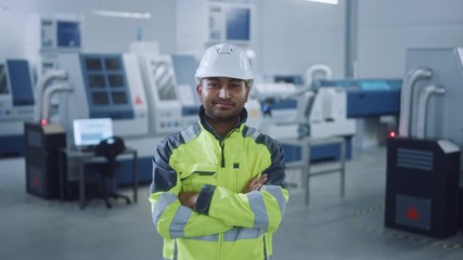 Poster - Portrait of Handsome Indian Engineer Wearing Safety Vest and Hardhat Crosses Arms and Smiles. Professional Woman Working in the Modern Manufacturing Factory. Facility with CNC Machinery and robot arm