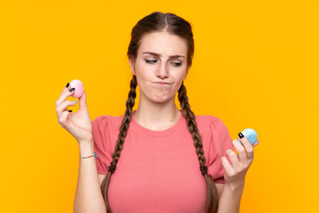 Wall Mural - Young woman over isolated yellow background holding colorful French macarons and unhappy