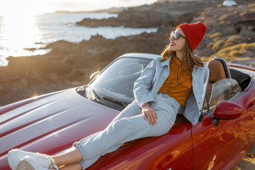 Young stylish woman enjoying beautiful landscapes, lying on the car hood while travel on the rocky coastline on a sunset. Carefree lifestyle and car travel concept