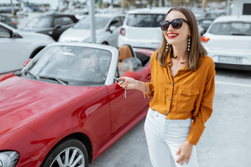 Portrait of a beautiful young woman standing with keys near the red cabriolet at the car parking outdoors. Concept of a happy car buying or renting