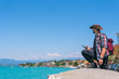 © Sergey - A man with smartpone dressed in a cowboy hat and a backpack sits on the promenade, looks at the turquoise water of Lake Garda. Vacation. Travels and tours to Europe. Sunny summer day. Italy