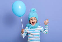 Horizontal Studio Picture Of Joyful Sweet Emotional Little Girl Opening Her Mouth And Mouth Widely, Raising Arms, Holding Blue Balloon, Enjoying Her Free Time, Playing Games. Childhood Concept.