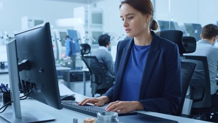 Modern Factory Office: Portrait of Young and Confident Female Industrial Engineer Working on Computer Holding Metal Component and Inspecting it. Big Industrial Gigafactory with High Tech CNC Machinery
