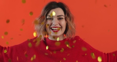 Wall Mural - Portrait of young sexy girl blowing shiny gold confetti with her hands in front herself and smiling looking camera on red background festive party. Falling sweets close up. Glitter. Monotone. Emotions