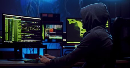 View from the side on the man in a hood working as a hacker at the computer in the dark room at night, hacking the system and laundring secret information.