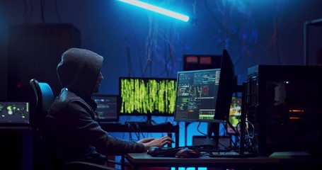 Side view on the Caucasian anonymous male cyber criminal in hood hacking data and coding viruses at the computer in the dark at night.
