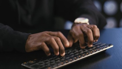 Worried-looking african-american mature man computer hacker wearing black hoodie is sitting in tech room and actively typing something on keyboard, staring at display in dark room.