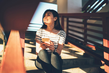 Wall Mural - Positive asian female blogger enjoying free time feeling happy about sun rays while sitting on stairs, cheerful young hipster girl satisfied with getting new updates for mobile phone applications.