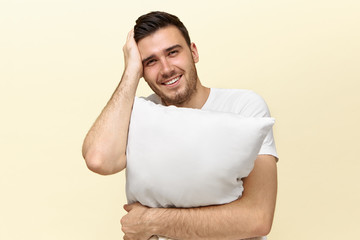 Wall Mural - Studio image of handsome tired young Caucasian man looking at camera with happy smile needs some sleep because of stressful day at work, holding white orthopedic pillow, keeping hand on face