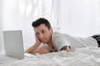 © ianachyrva - Young man in the white shirt on a bed with white linen is excited looking at the screen of a laptop.