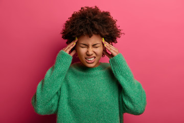 Wall Mural - Upset dark skinned curly woman touches temples, frowns face and suffers from migraine, terrible headache, needs painkillers, wears warm green sweater, closes eyes to relieve pain, poses indoor