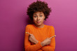 Unaware hesitant woman with curly hairstyle crosses hands over chest, points sideways, feels confused while chooses between two objects, wears orange casual jumper, models over purple studio wall