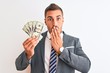 © Krakenimages.com - Young handsome business man holding bunch of dollars banknotes over isolated background cover mouth with hand shocked with shame for mistake, expression of fear, scared in silence, secret concept