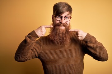 Canvas Print - Handsome Irish redhead man with beard wearing glasses and winter sweater over yellow background smiling cheerful showing and pointing with fingers teeth and mouth. Dental health concept.