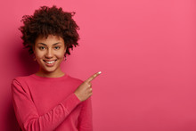 Pleasant Looking Cheerful Afro American Woman Points At Upper Right Corner, Discusses Advertisement, Gives Advice What To Buy, Smiles Gently, Wears Pink Jumper. Find Best Price And Discount There
