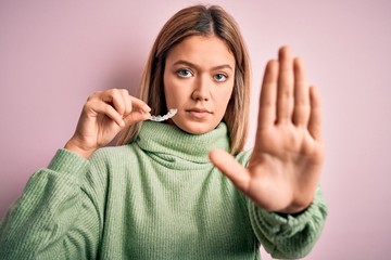 Sticker - Young beautiful woman holding aligner standing over isolated pink background with open hand doing stop sign with serious and confident expression, defense gesture