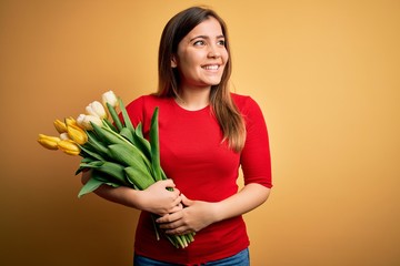 Canvas Print - Young blonde woman holding romantic bouquet of tulips flowers over yellow background looking away to side with smile on face, natural expression. Laughing confident.