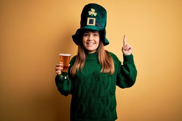 Sticker - Young beautiful woman wearing green hat drinking glass of beer on saint patricks day smiling amazed and surprised and pointing up with fingers and raised arms.