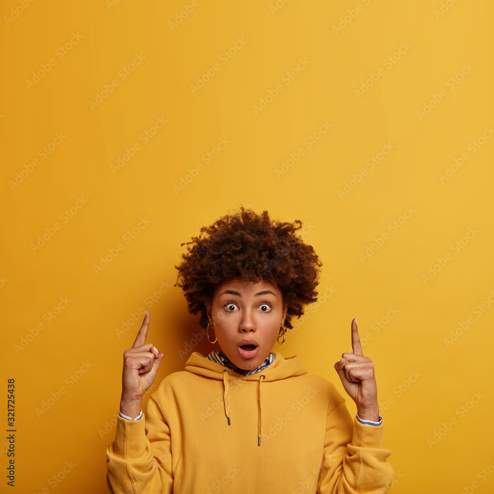 Wow, look there! Impressed shocked woman with Afro hair points fore fingers above, shows blank space, stunned by very high price, shows place for your advertisement, isolated over yellow background - obrazy, fototapety, plakaty 