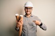 Young architect man wearing contractor helmet holding project paper plan over isolated background with surprise face pointing finger to himself