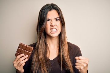 Poster - Young beautiful girl holding sweet bar of chocolate over isolated white background annoyed and frustrated shouting with anger, crazy and yelling with raised hand, anger concept