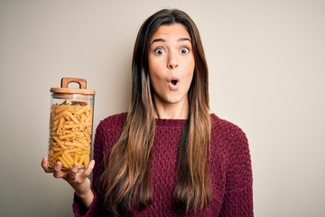 Young beautiful girl holding bottle of dry Italian pasta macaroni over white background scared in shock with a surprise face, afraid and excited with fear expression