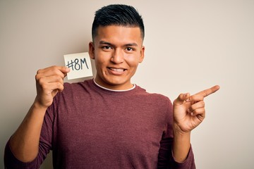 Young handsome latin man celebrating 8th march womens day holding reminder paper very happy pointing with hand and finger to the side