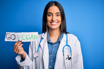 Young beautiful brunette doctor woman wearing coat holding paper with go vegan message with a happy face standing and smiling with a confident smile showing teeth