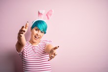 Young Woman With Fashion Blue Hair Wearing Easter Rabbit Ears Over Pink Background Looking At The Camera Smiling With Open Arms For Hug. Cheerful Expression Embracing Happiness.