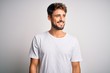 © Krakenimages.com - Young handsome man with beard wearing casual t-shirt standing over white background looking away to side with smile on face, natural expression. Laughing confident.