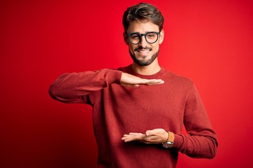 Wall Mural - Young handsome man with beard wearing glasses and sweater standing over red background gesturing with hands showing big and large size sign, measure symbol. Smiling looking at the camera. Measuring