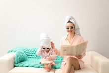 Mother And Little Daughter In Bathrobes With Newspapers Sitting On Sofa