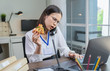 © sataporn - Young Asian businesswoman having pizza snack while talking phone with customers due to working late.