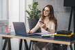 © foxyburrow - Female executive sitting at desk in office, using portable digital devices for work. Girl typing on laptop and texting on smartphone. Young woman taking selfie at workplace by computer on camera