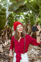 Wall Mural - Woman as a tourist dressed in red exploring banana plantation, photographing or vlogging on phone. Concept of a healthy eating or green tourism